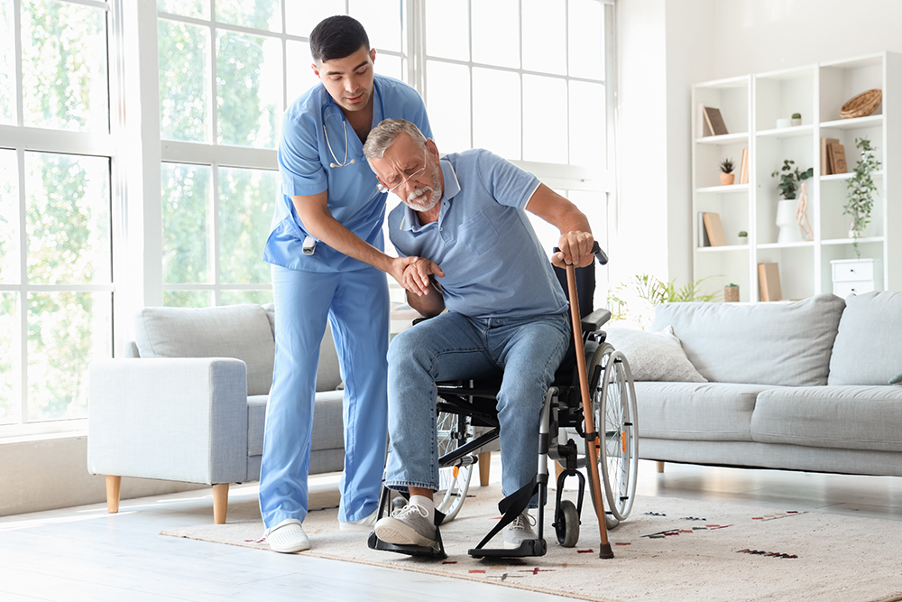 Younger male certified home health aide assisting older male client with getting out of a wheelchair