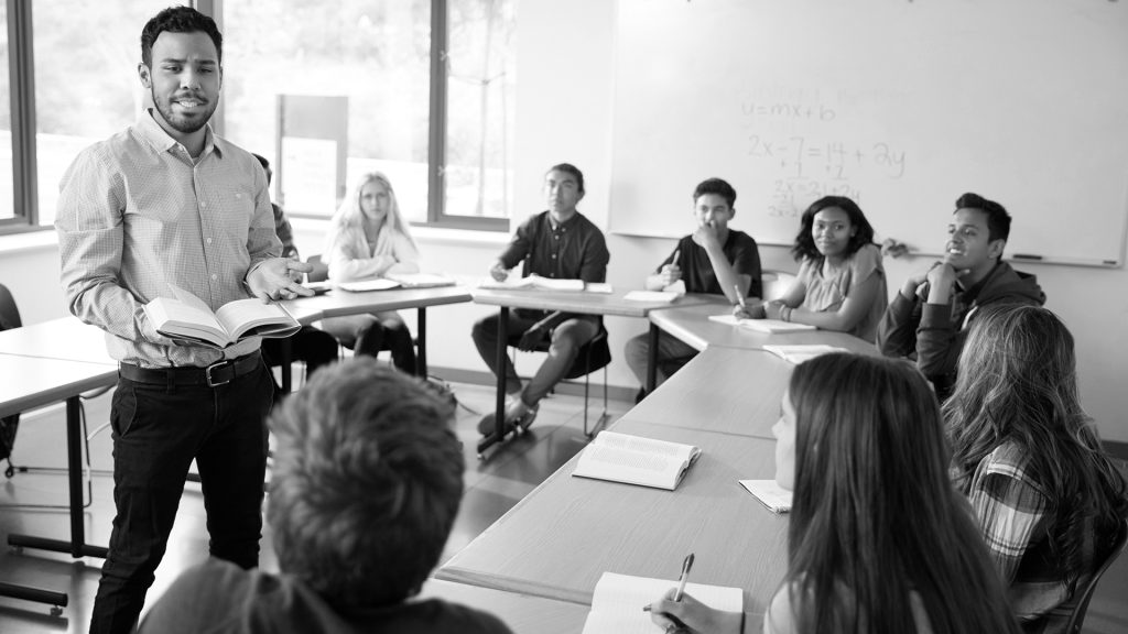 A teacher holding a book with 8 students sitting in a round table configuration